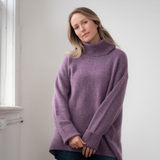 Woman modelling an oversized purple cowl neck sweater with blue jeans in a white room photographed near a window