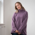 Woman modelling an oversized purple cowl neck sweater with blue jeans in a white room photographed near a window