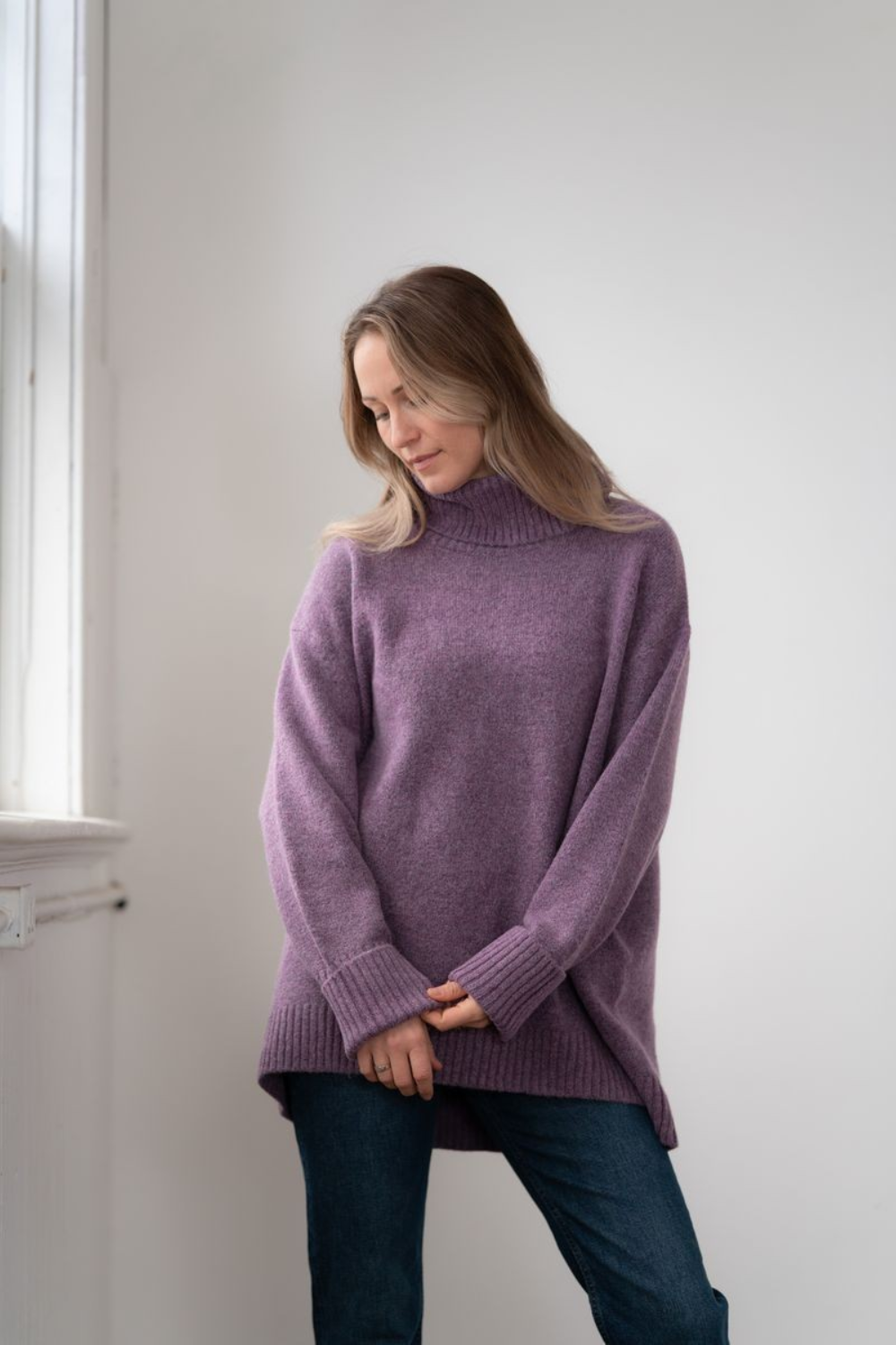 Woman modelling an oversized purple cowl neck sweater with blue jeans in a white room photographed near a window