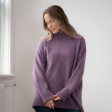 Woman modelling an oversized purple cowl neck sweater with blue jeans in a white room photographed near a window
