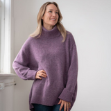 Woman modelling an oversized purple cowl neck sweater with blue jeans in a white room photographed near a window