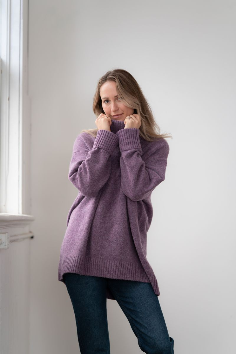 Woman modelling an oversized purple cowl neck sweater with blue jeans in a white room photographed near a window