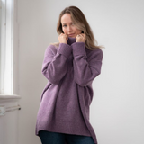 Woman modelling an oversized purple cowl neck sweater with blue jeans in a white room photographed near a window