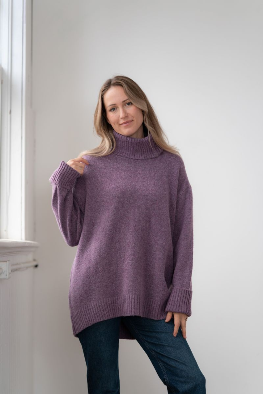Woman modelling an oversized purple cowl neck sweater with blue jeans in a white room photographed near a window