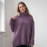 Woman modelling an oversized purple cowl neck sweater with blue jeans in a white room photographed near a window