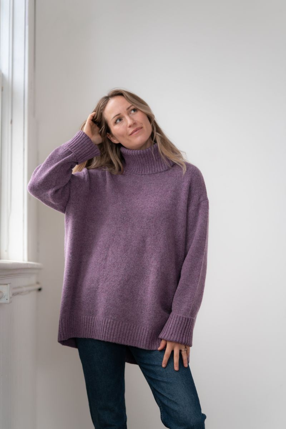 Woman modelling an oversized purple cowl neck sweater with blue jeans in a white room photographed near a window