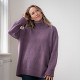 Woman modelling an oversized purple cowl neck sweater with blue jeans in a white room photographed near a window