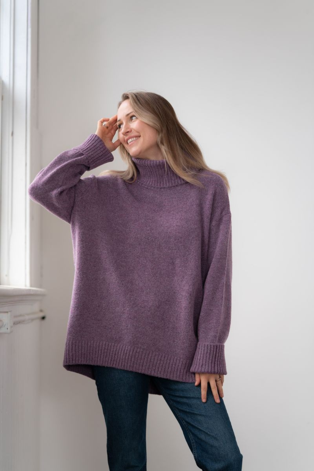 Woman modelling an oversized purple cowl neck sweater with blue jeans in a white room photographed near a window