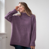 Woman modelling an oversized purple cowl neck sweater with blue jeans in a white room photographed near a window