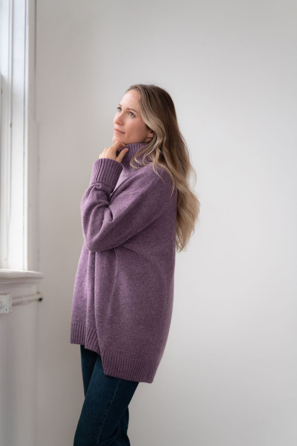 Woman modelling an oversized purple cowl neck sweater with blue jeans in a white room photographed near a window