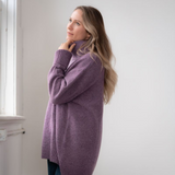 Woman modelling an oversized purple cowl neck sweater with blue jeans in a white room photographed near a window