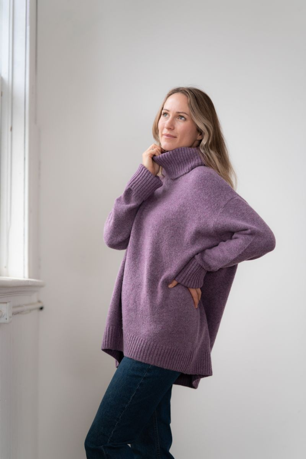 Woman modelling an oversized purple cowl neck sweater with blue jeans in a white room photographed near a window