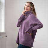 Woman modelling an oversized purple cowl neck sweater with blue jeans in a white room photographed near a window