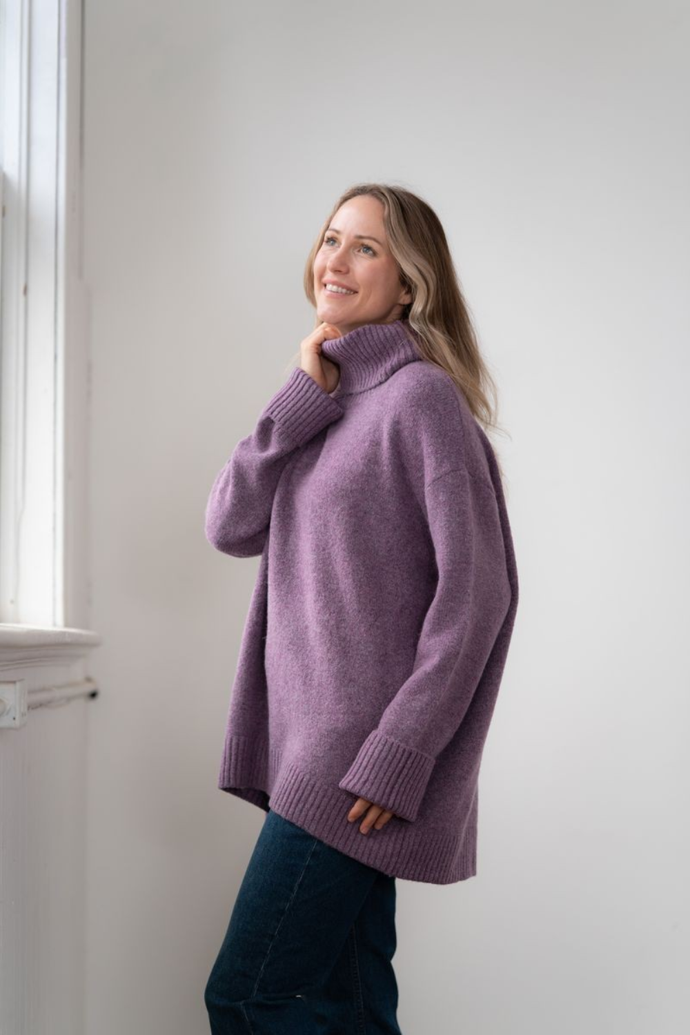 Woman modelling an oversized purple cowl neck sweater with blue jeans in a white room photographed near a window
