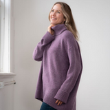 Woman modelling an oversized purple cowl neck sweater with blue jeans in a white room photographed near a window