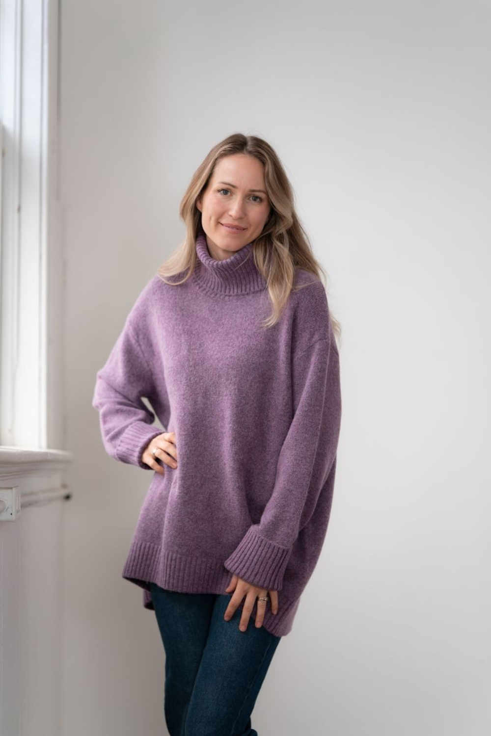 Woman modelling an oversized purple cowl neck sweater with blue jeans in a white room photographed near a window