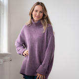 Woman modelling an oversized purple cowl neck sweater with blue jeans in a white room photographed near a window