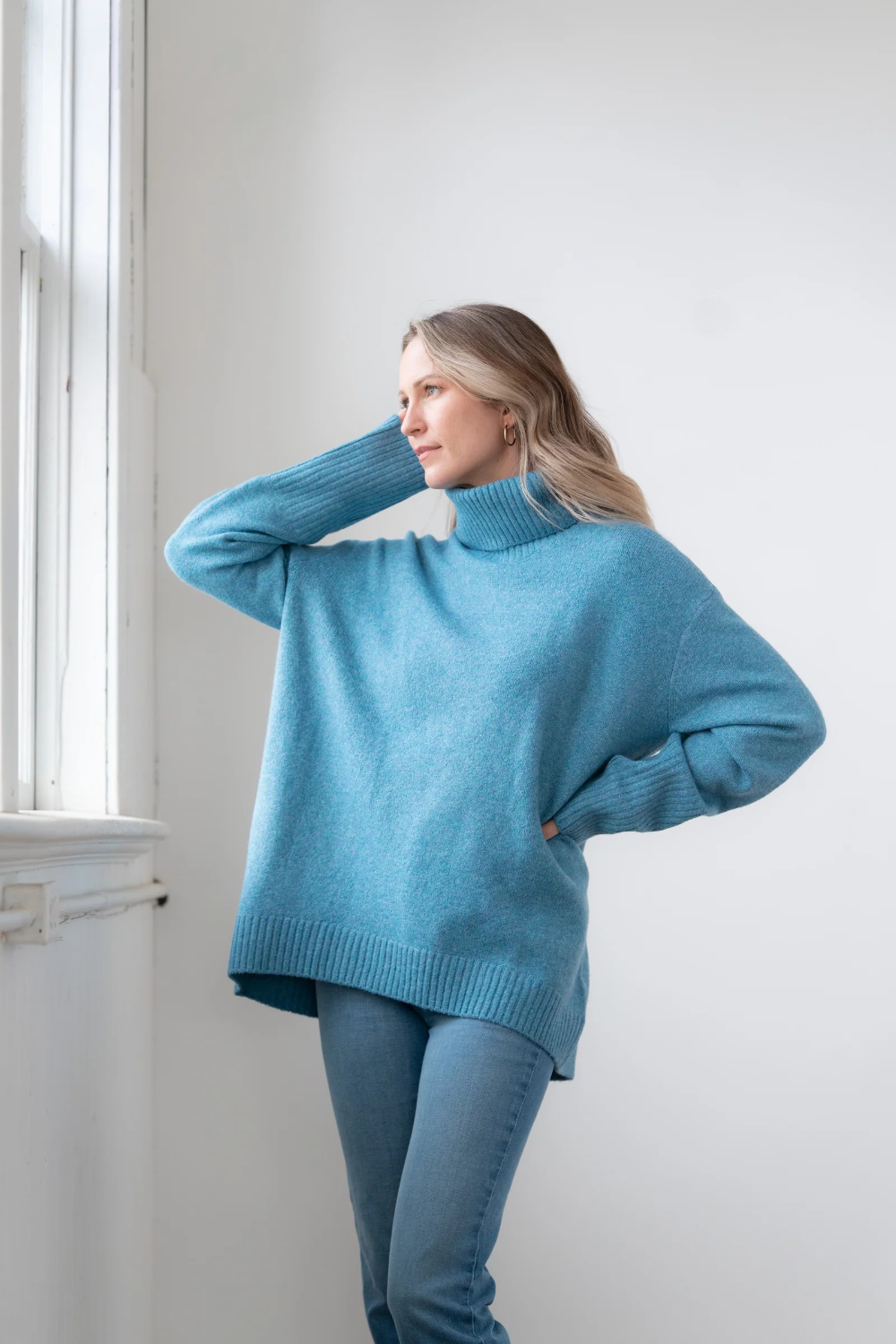 Woman modelling an oversized light blue chunky turtle neck sweater with blue jeans standing indoors, near a window