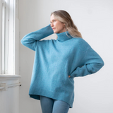 Woman modelling an oversized light blue chunky turtle neck sweater with blue jeans standing indoors, near a window