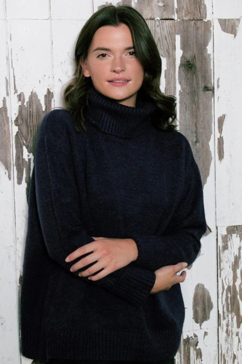 Woman wearing over sized black turtle neck sweater standing in front of a distressed wood wall.