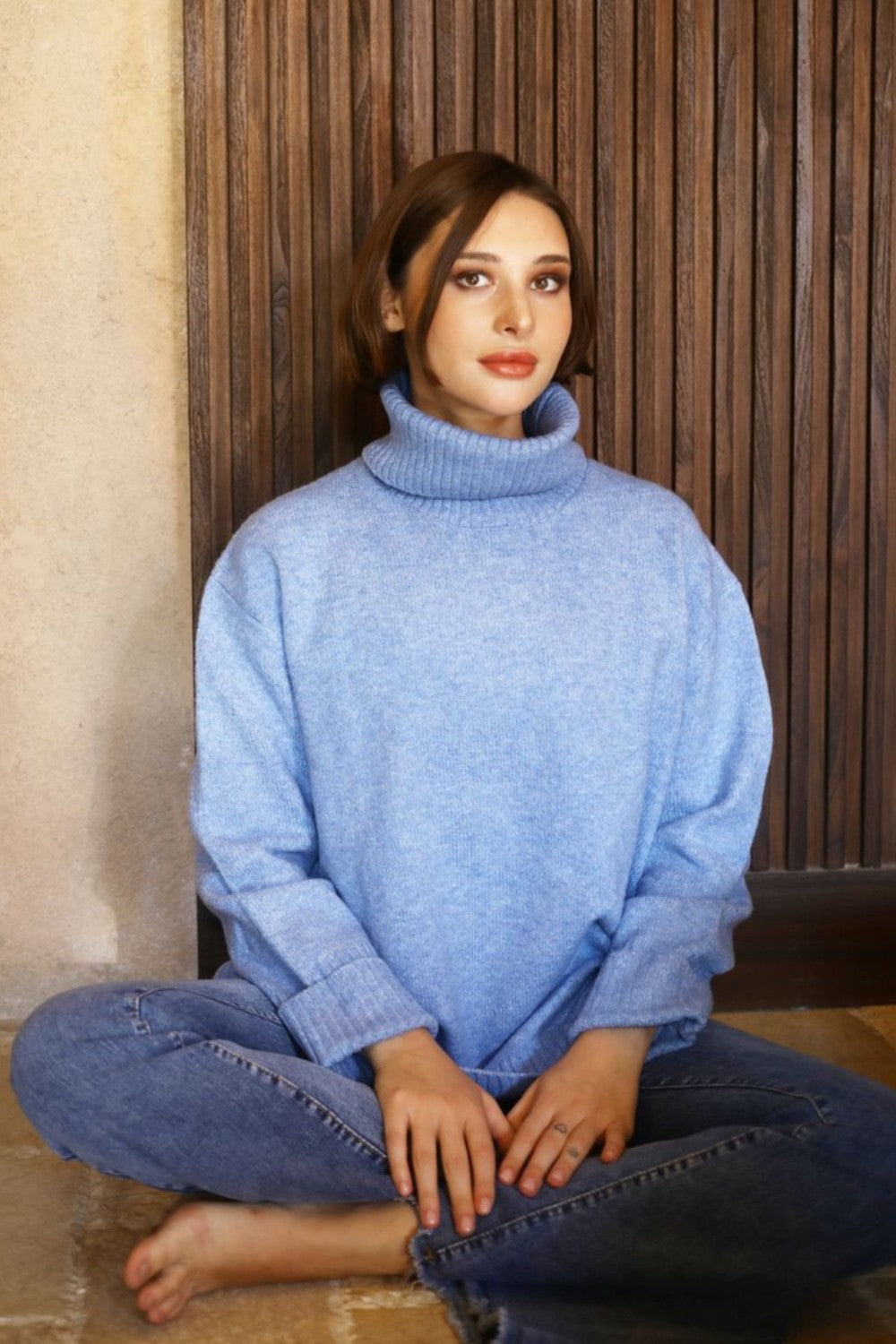 Woman sitting cross legged on the floor wearing a blue chunky turtle neck sweater and jeans. 