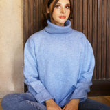 Woman sitting cross legged on the floor wearing a blue chunky turtle neck sweater and jeans. 