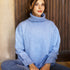 Woman sitting cross legged on the floor wearing a blue chunky turtle neck sweater and jeans. 