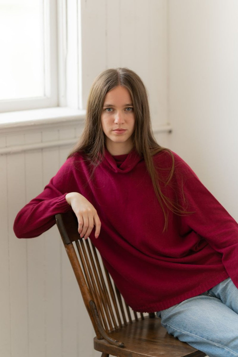 Woman sitting on a wood chair wearing loose red sweater and blue jeans.