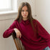 Woman sitting on a wood chair wearing loose red sweater and blue jeans.