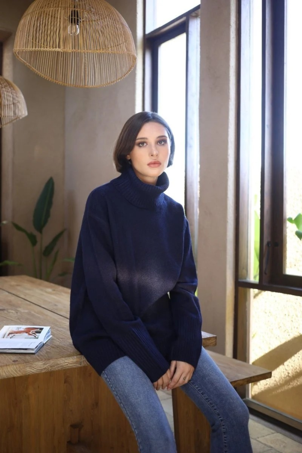 Woman sitting indoors, near a window, wearing an oversized navy chunky turtle neck sweater with blue jeans