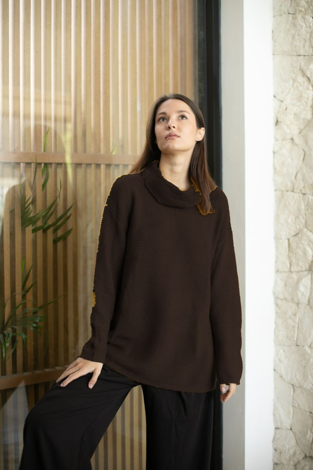 A woman wearing a dark brown ribbed knit sweater stands in front of a window next to a stone wall.