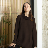 A woman wearing a dark brown ribbed knit sweater stands in front of a window next to a stone wall.