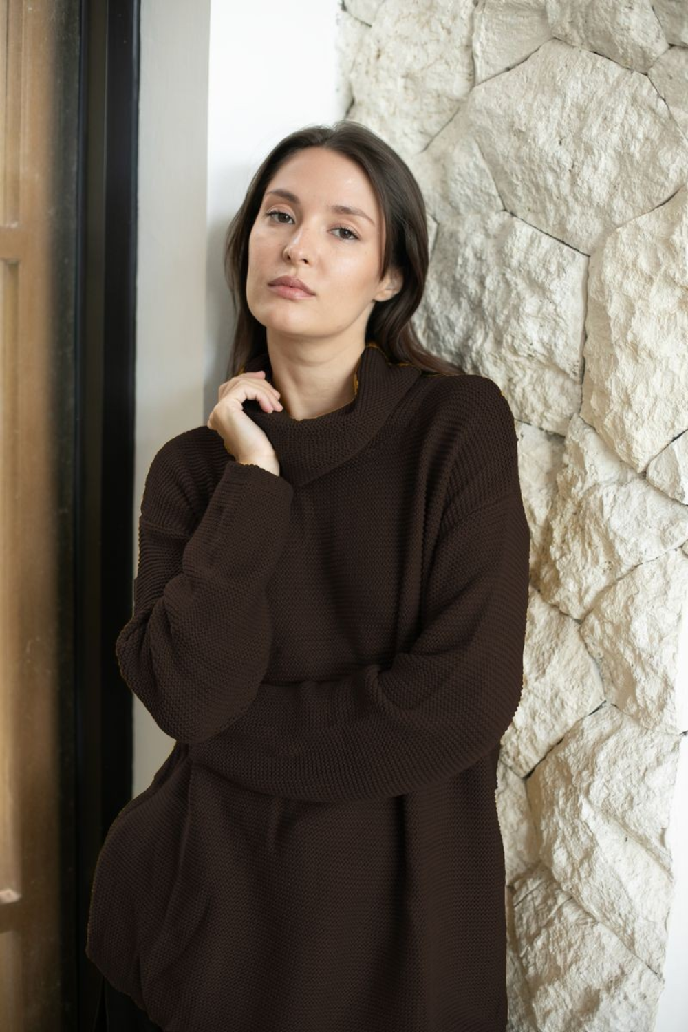 A woman wearing a dark brown ribbed knit sweater leans against a textured stone wall next to a doorway.