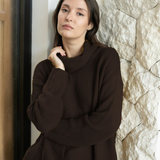 A woman wearing a dark brown ribbed knit sweater leans against a textured stone wall next to a doorway.