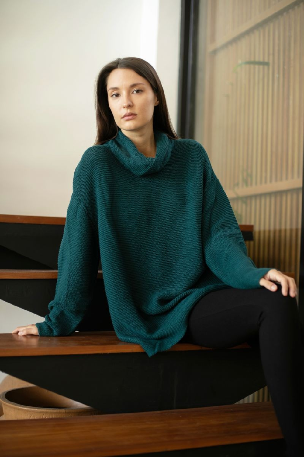 A woman wearing an oversized dark teal ribbed knit cowl-neck sweater and black leggings, sitting on a modern wooden staircase indoors.
