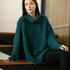 A woman wearing an oversized dark teal ribbed knit cowl-neck sweater and black leggings, sitting on a modern wooden staircase indoors.