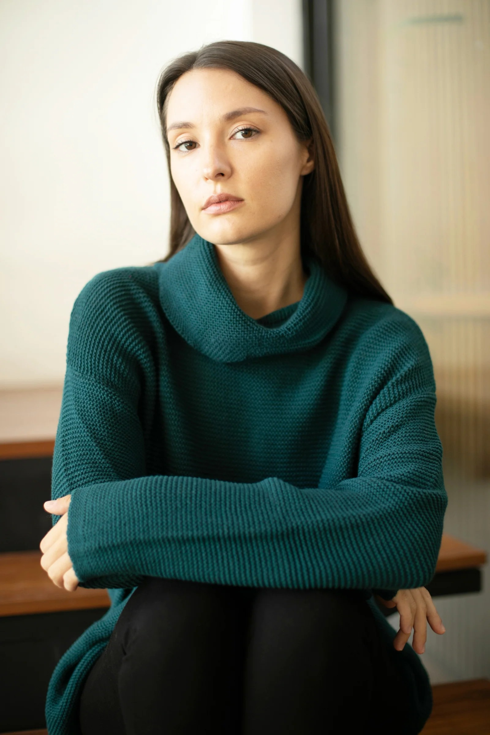 A woman with long dark brown hair sits with her arms crossed, wearing a ribbed knit cowl-neck sweater in a deep teal green color, paired with black pants.