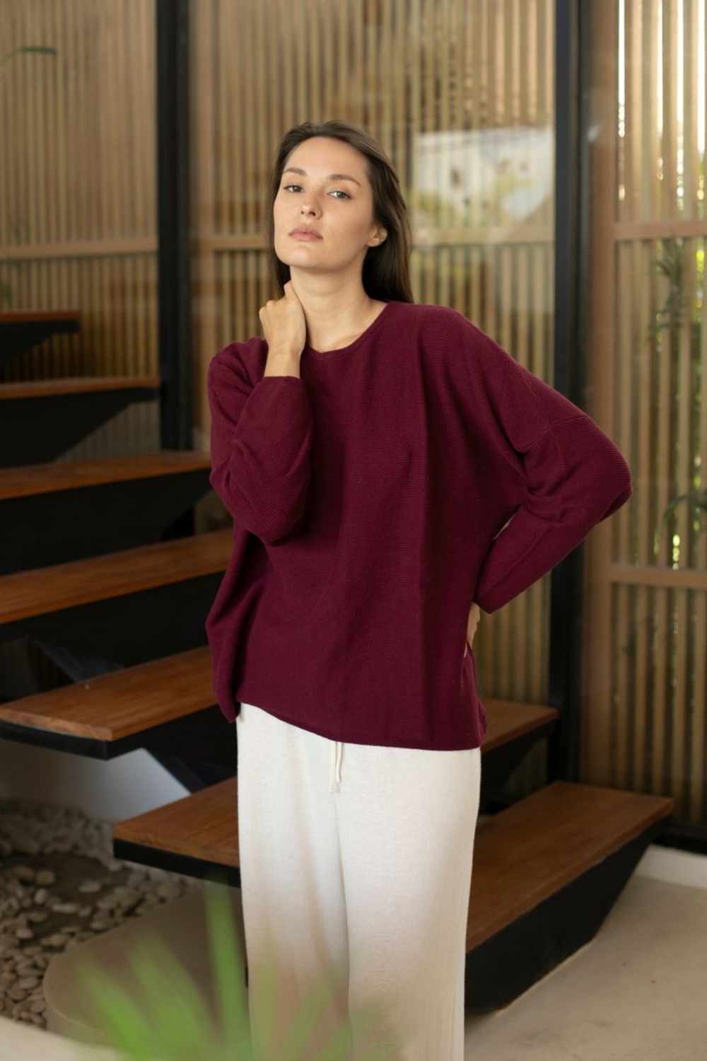A woman wearing a loose-fitting, long-sleeve burgundy knit top and white pants, standing in a modern interior with wooden steps and slatted walls.