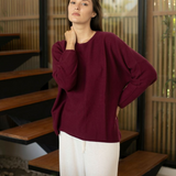 A woman wearing a loose-fitting, long-sleeve burgundy knit top and white pants, standing in a modern interior with wooden steps and slatted walls.