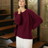 A woman wearing a loose-fitting, long-sleeve burgundy knit top and white pants, standing in a modern interior with wooden steps and slatted walls.