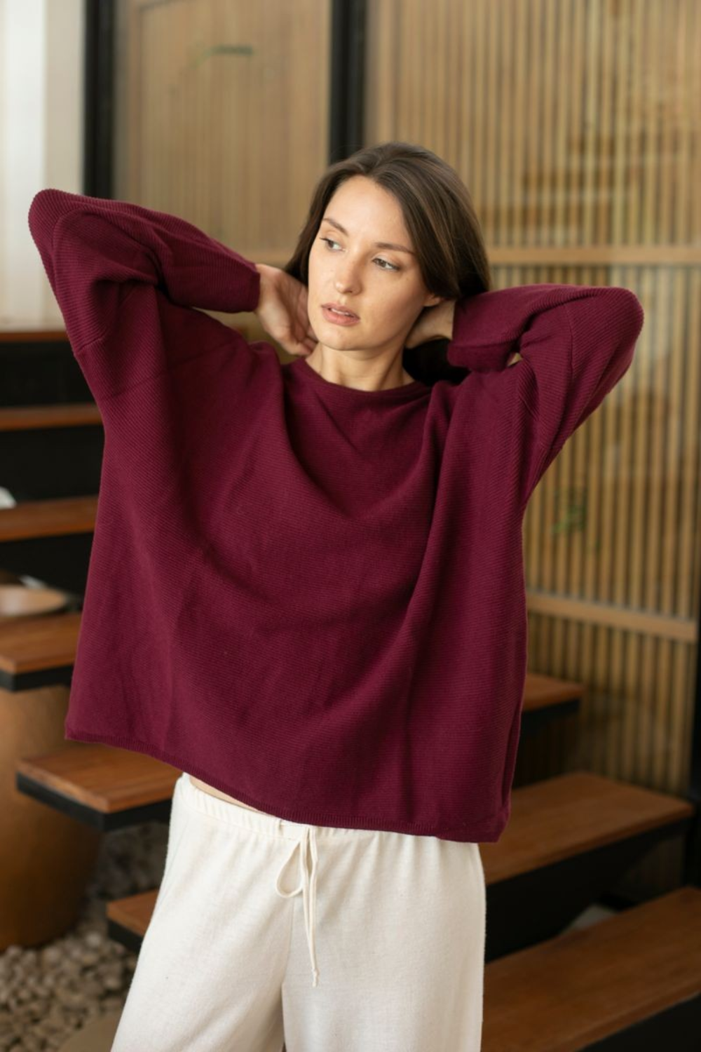 A woman wearing a loose, oversized crewneck sweater in deep burgundy and white drawstring pants, posing with her hands behind her head in an indoor setting with wooden screens.