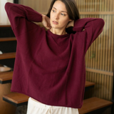 A woman wearing a loose, oversized crewneck sweater in deep burgundy and white drawstring pants, posing with her hands behind her head in an indoor setting with wooden screens.