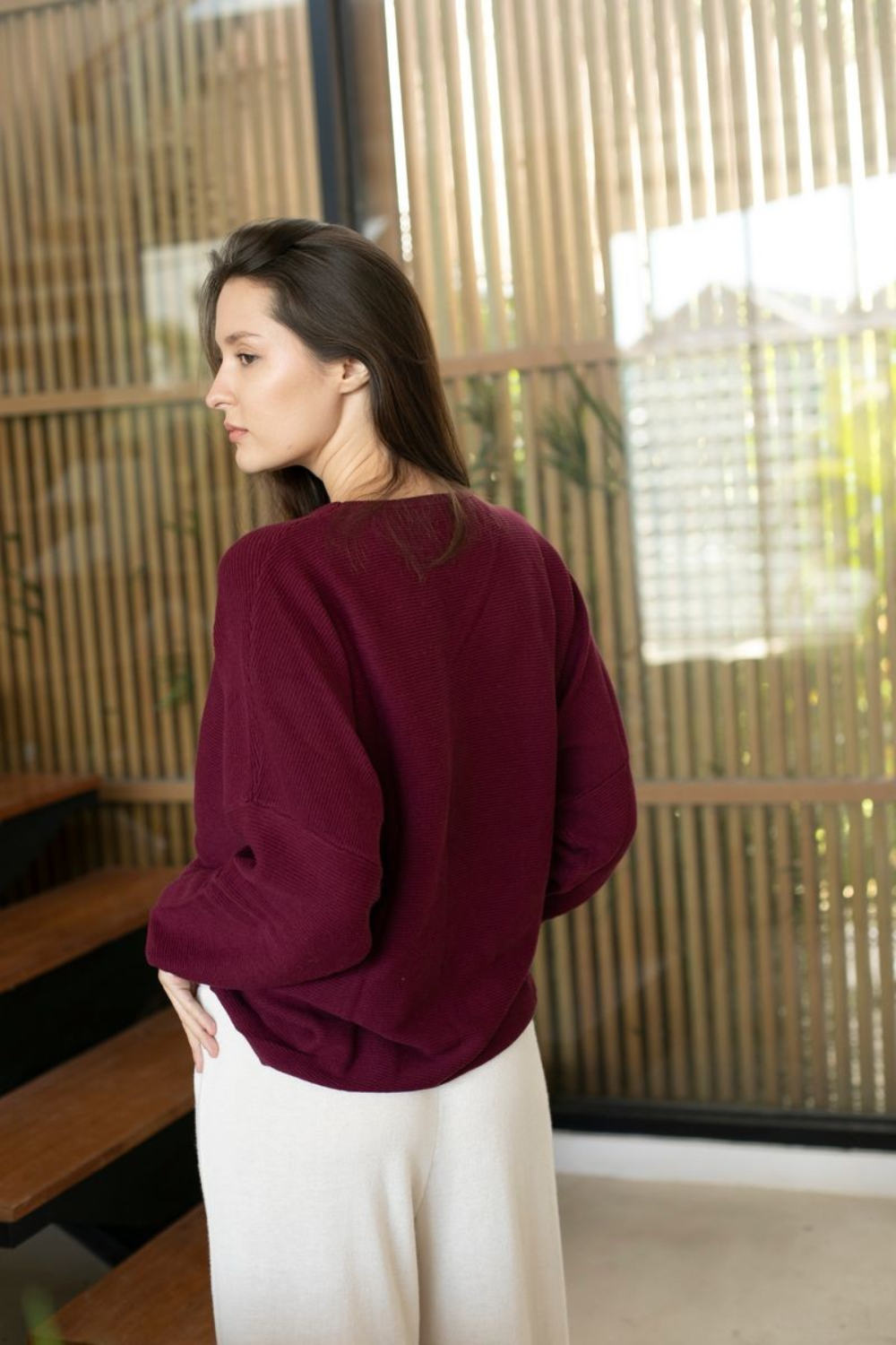 A woman wearing a deep burgundy long-sleeve top and white pants, standing in profile in front of a modern wooden slatted screen.