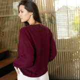 A woman wearing a deep burgundy long-sleeve top and white pants, standing in profile in front of a modern wooden slatted screen.
