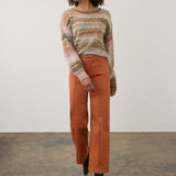 A woman stands in a studio wearing a fuzzy striped knit sweater in muted pink, beige, and brown tones, paired with rust-orange wide-leg pants and dark brown ankle boots against a plain off-white wall and concrete floor.