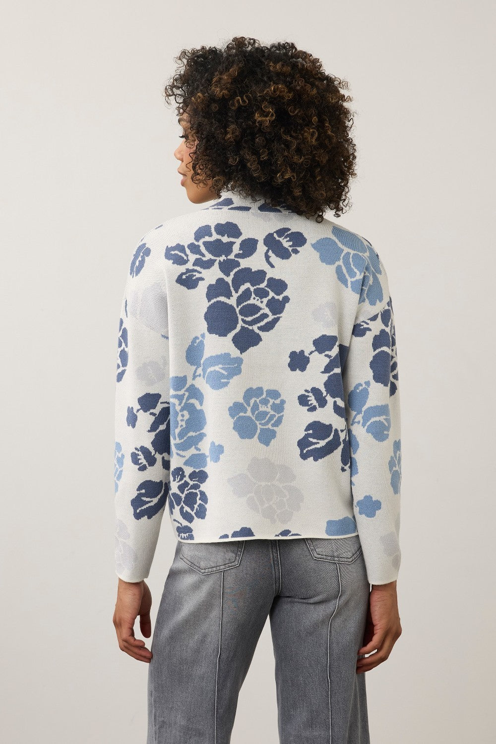 A woman viewed from the back wearing a white knit cardigan with a blue and gray floral pattern, paired with gray jeans, against a plain light gray studio background.