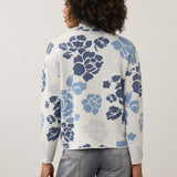 A woman viewed from the back wearing a white knit cardigan with a blue and gray floral pattern, paired with gray jeans, against a plain light gray studio background.