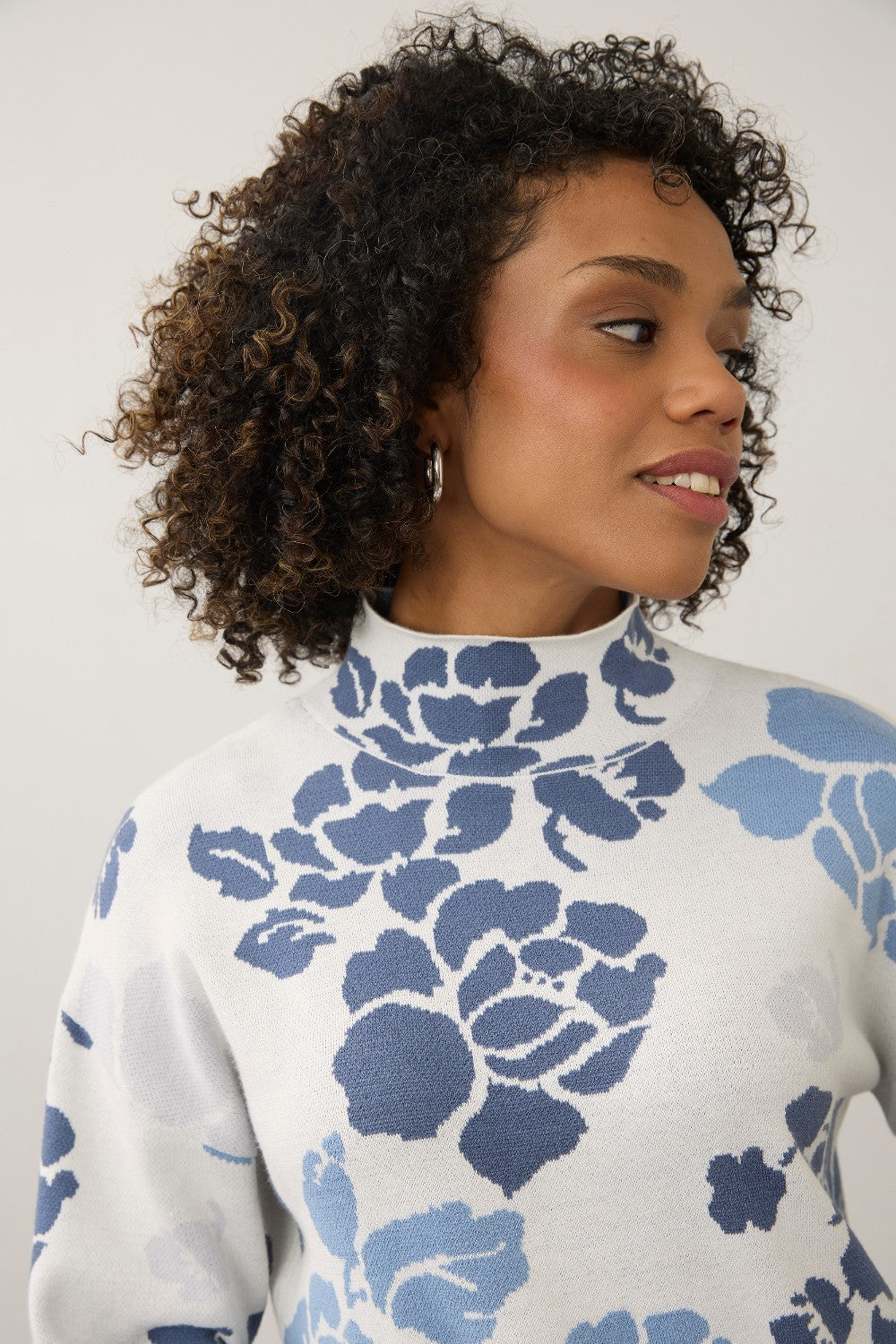 A close-up of a woman with curly brown hair wearing a white mock-neck knit sweater with a blue floral pattern, set against a plain light gray background.