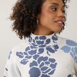 A close-up of a woman with curly brown hair wearing a white mock-neck knit sweater with a blue floral pattern, set against a plain light gray background.