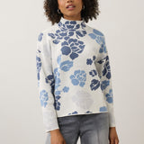 A woman with curly dark hair wearing a white mock-neck long-sleeve top with a blue floral pattern, paired with gray jeans, standing against a light gray studio background.